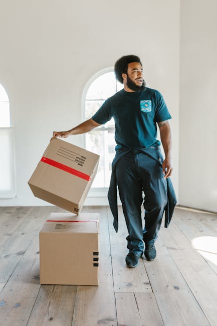 A man with dark curly hair and a beard, dressed in casual navy clothing, is standing in a bright, empty room with white walls and natural light coming through a large arched window. He is holding a cardboard moving box wrapped with red packing tape in his right hand, with two additional similarly packed boxes placed on the wooden floor beside him. The scene depicts a home relocation or packing process, possibly as part of a furniture transport or moving service. The room appears to be part of a residential property, and the natural lighting emphasizes the clean, minimal environment. Wandsworth Man and Van’s professional house removals services may involve packing, loading, and transporting household items, as suggested by this scene of handling boxes in a spacious interior.