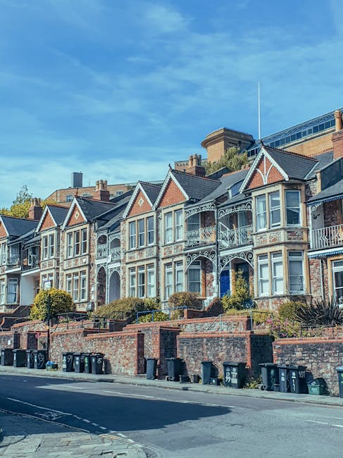 A row of Victorian-style terraced houses located on a residential street in Clapham Junction, Wandsworth, with detailed brick facades, ornate white ironwork balconies, and bay windows. The houses are built on a slight incline, with brick retaining walls and small front gardens containing neatly trimmed bushes. The street in front features a paved road with several black wheelie bins lined along the curb. The sky above is clear and blue, indicating daytime. This image illustrates a typical urban neighbourhood suitable for residential home relocation and furniture transport, with the houses providing context for moving or packing services in Wandsworth. Wandsworth Man and Van, a professional removals provider, often assist with these types of house moves, ensuring safe packing and loading processes in such property environments.