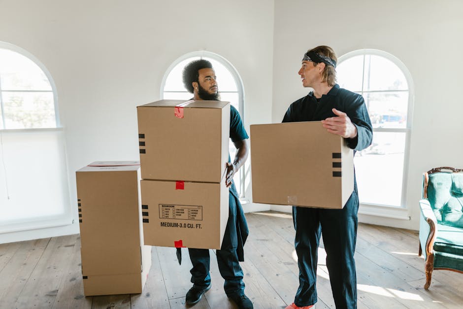 Two movers from Wandsworth Man and Van are inside a well-lit room with large, arched windows, engaged in a home relocation process. The mover on the left, an African-American man with curly hair and a beard, is holding a medium-sized cardboard box, with a stack of three similar boxes in front of him on the wooden floor. The middle box has red packing tape and labels indicating it is a medium three cubic foot carton. The second mover, a Caucasian man with long blond hair wearing a headband and dark clothing, is carrying an identical cardboard box, gesturing as he converses with the other mover. The room has a neutral-colored wall and minimal furnishings, including a green upholstered armchair partially visible on the right side, suggesting the ongoing packing and moving activities associated with furniture transport and household packing. The scene captures the loading process during a house move, emphasizing careful handling of packed boxes by professional removals service, suitable for the SW18 moving and removals guide for Wandsworth.