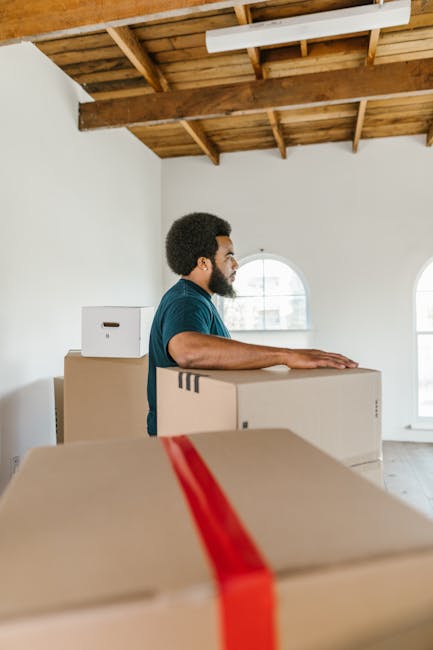 A man with dark curly hair and a beard, dressed in a teal shirt, is standing inside a spacious, well-lit room with a high wooden ceiling and exposed beams. He is next to several packed cardboard moving boxes, some of which are sealed with red packing tape, indicating they are ready for a home relocation. The boxes are of various sizes, with one directly in front of him and another stacked behind. The room has white walls and two large arched windows, allowing natural light to illuminate the scene. In the background, there is no visible furniture or appliances, but the presence of numerous boxes suggests ongoing packing and moving preparations, typical of furniture transport and packing activities associated with professional removals. This setup reflects the early stages of a house moving process, coordinated by Wandsworth Man and Van, specializing in removals in the SW18 area, facilitating efficient loading and packing for a smooth instructions for home relocation.