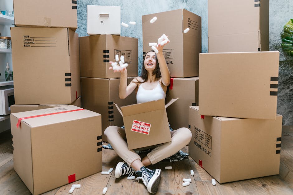Two movers from Wandsworth Man and Van are inside a well-lit room with large, arched windows, engaged in a home relocation process. The mover on the left, an African-American man with curly hair and a beard, is holding a medium-sized cardboard box, with a stack of three similar boxes in front of him on the wooden floor. The middle box has red packing tape and labels indicating it is a medium three cubic foot carton. The second mover, a Caucasian man with long blond hair wearing a headband and dark clothing, is carrying an identical cardboard box, gesturing as he converses with the other mover. The room has a neutral-colored wall and minimal furnishings, including a green upholstered armchair partially visible on the right side, suggesting the ongoing packing and moving activities associated with furniture transport and household packing. The scene captures the loading process during a house move, emphasizing careful handling of packed boxes by professional removals service, suitable for the SW18 moving and removals guide for Wandsworth.